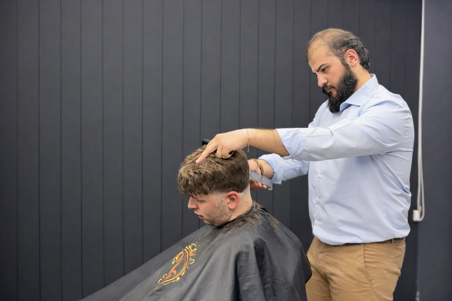 Barbeiro de óculos e luvas pretas finalizando o corte de um cliente em uma barbearia moderna e minimalista.